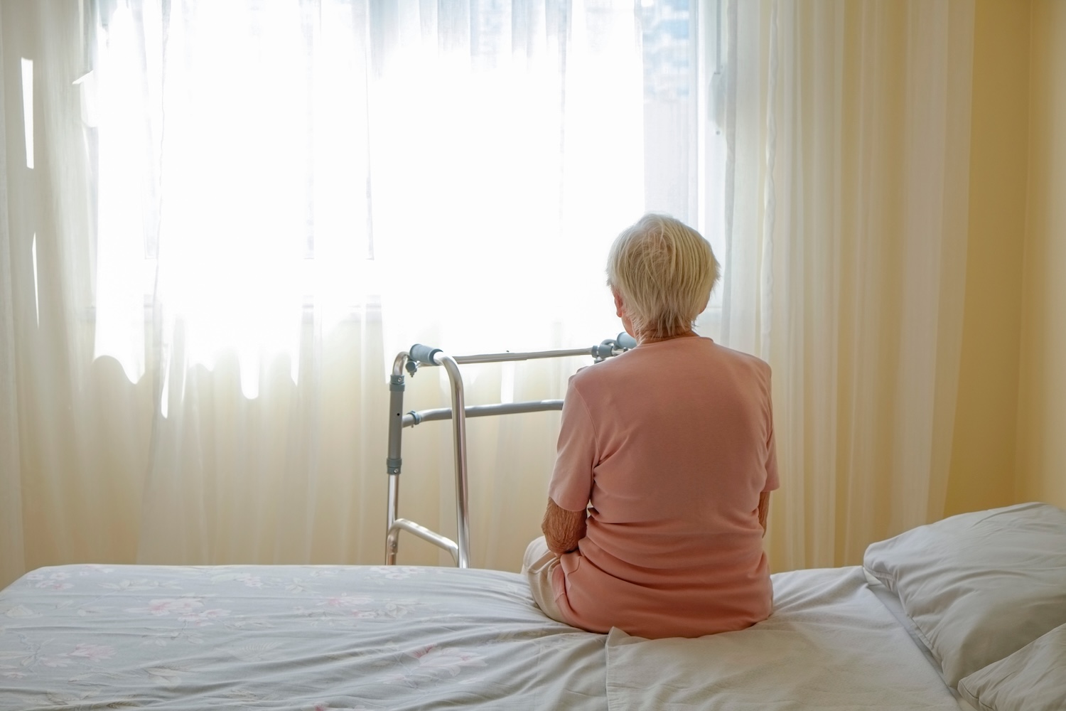 Older woman looking through curtains sitting on bed