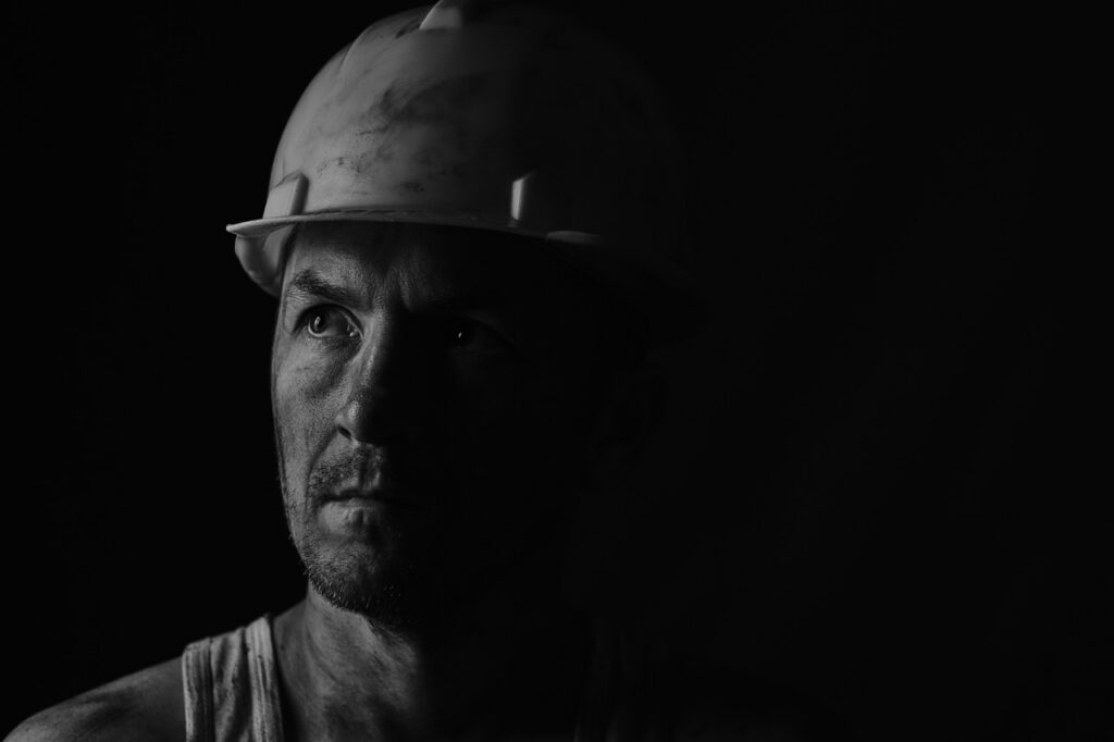 Black and white portrait of a construction worker wearing a hard hat, his face covered in dust in a dark setting, evoking concerns about workplace hazards such as asbestos exposure.